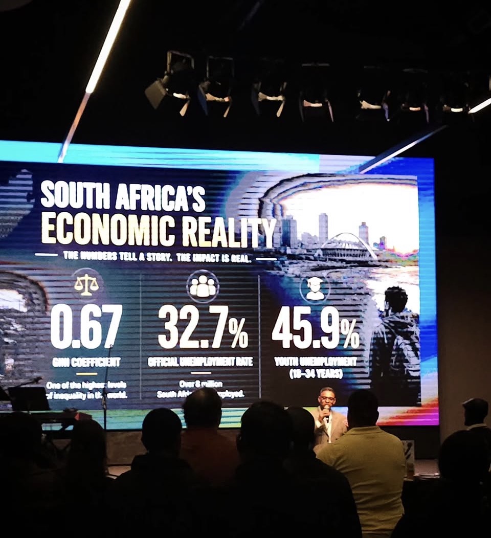 A presenter at the gala in front of a screen showing South Africa's economic reality